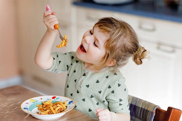 A young child with a bowl full of pasta with some on a fork smiling.